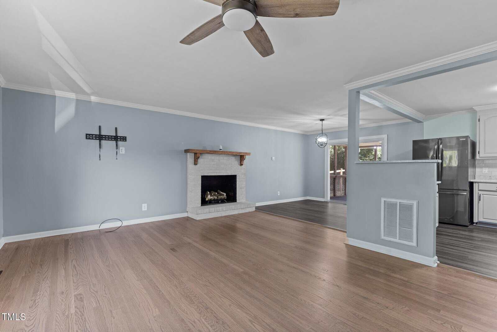 A spacious living room with light blue walls and wooden floors. There's a ceiling fan above and a fireplace with a wooden mantle. An open space leads to a kitchen area with white cabinets and stainless steel appliances. Natural light fills the room.