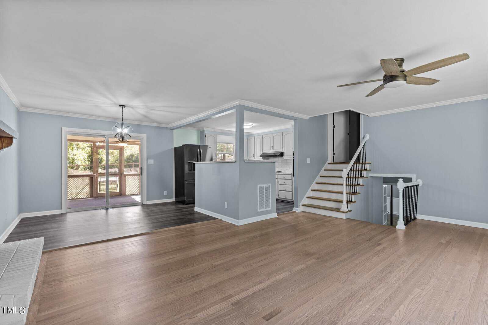 A spacious open-plan living room with light blue walls, hardwood floors, and a staircase leading upstairs. A ceiling fan is above, and sliding glass doors open to a wooden deck outside. The kitchen has a white countertop and stainless steel appliances.
