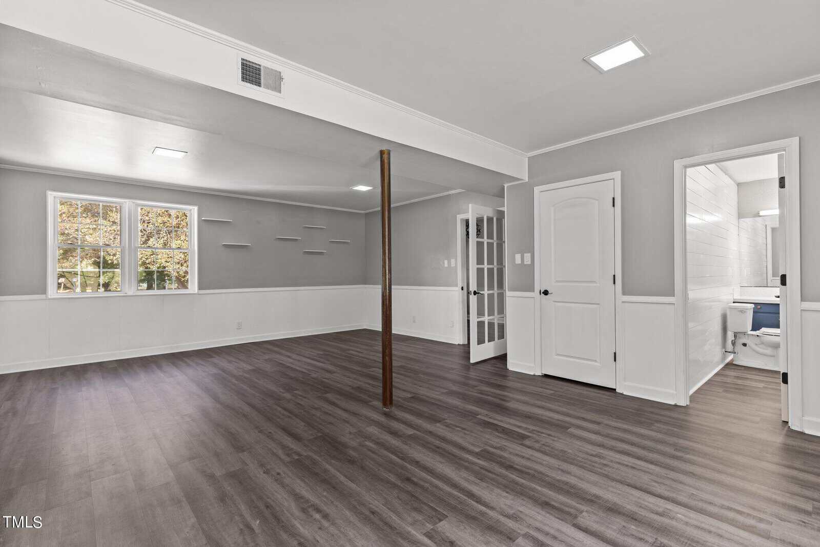 Spacious basement room with gray wood flooring, light gray walls, and recessed lighting. Features a wooden support beam, built-in shelves, a small window, and a doorway leading to a white-tiled bathroom with a bathtub.