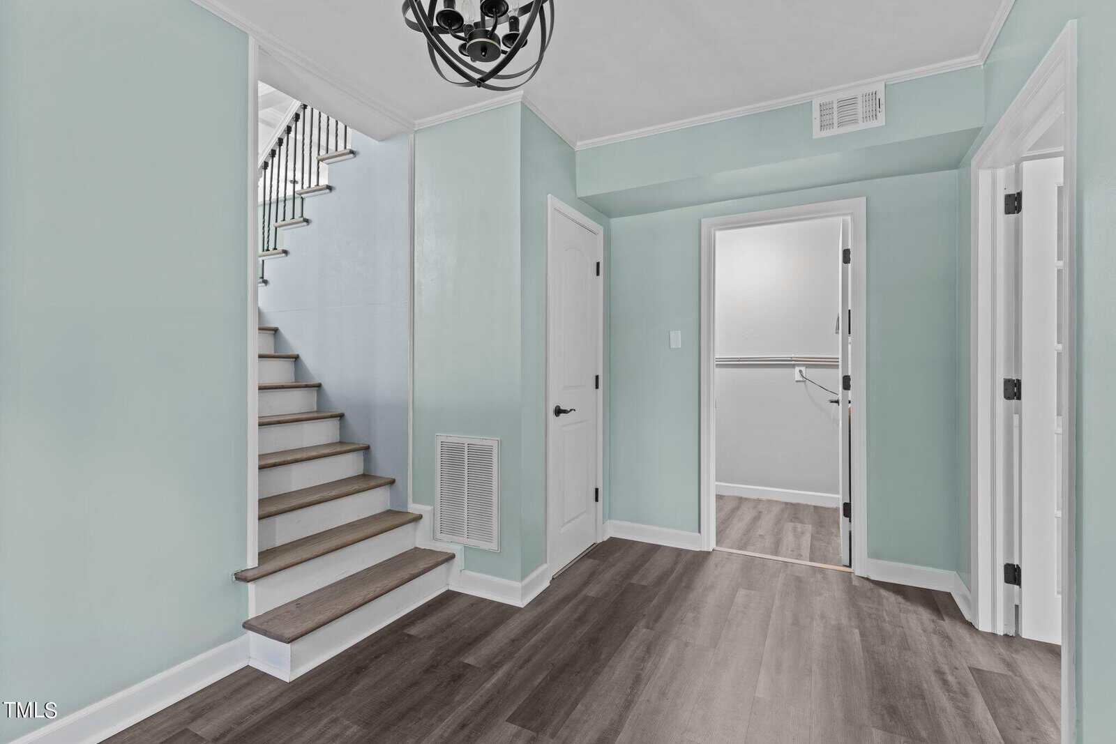 A hallway with light teal walls and wood-like flooring. There are three white doors, one leading to a small closet space. A wooden staircase with black railings is on the left. A black chandelier hangs from the ceiling.
