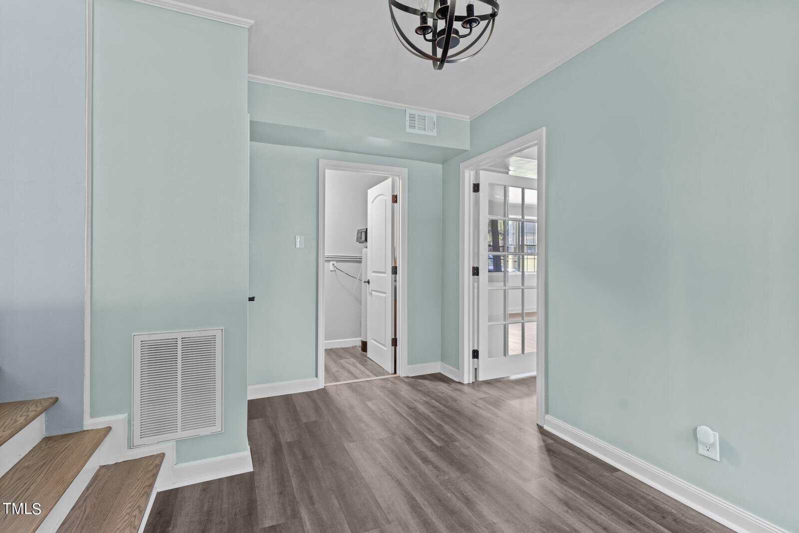 A small room with mint green walls and wooden flooring. There is a modern black chandelier hanging from the ceiling. A staircase on the left and a doorway leading to a closet are visible, along with a set of glass-paneled double doors on the right.