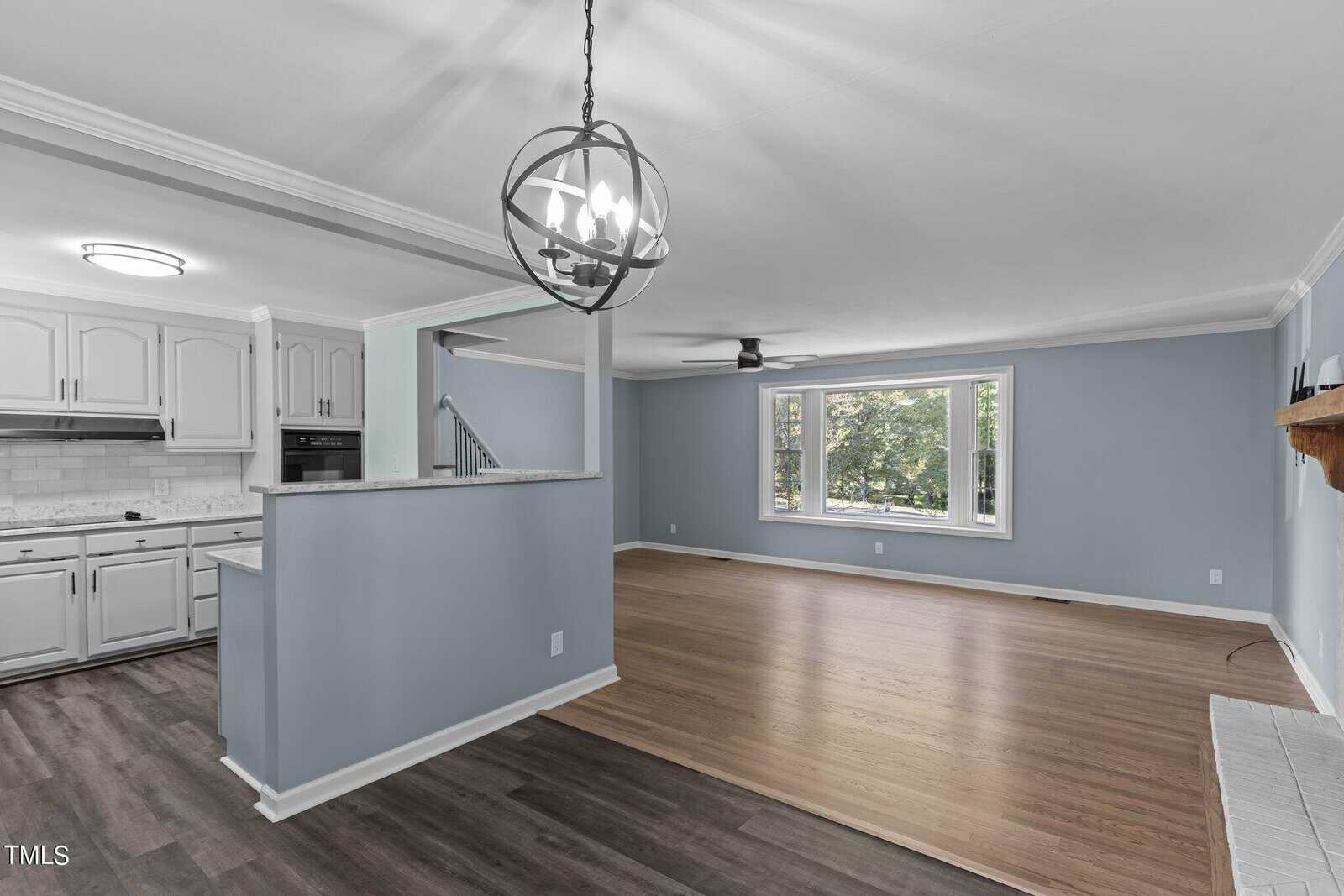 An open-concept living space with light blue walls, featuring a modern kitchen with white cabinets, a gray countertop, and a brick backsplash. The room has hardwood floors, a large window, and a spherical chandelier above the dining area.