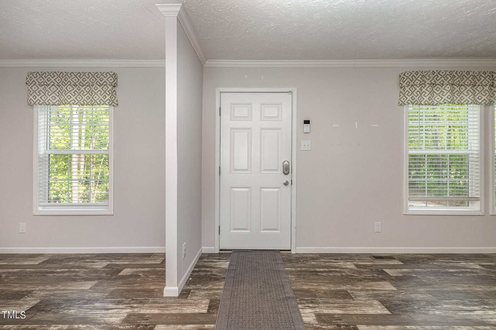 A bright entryway with a white front door centered between two windows. The walls are light grey, and the floor features a mix of wood and tile textures. Small patterned valances adorn the windows.