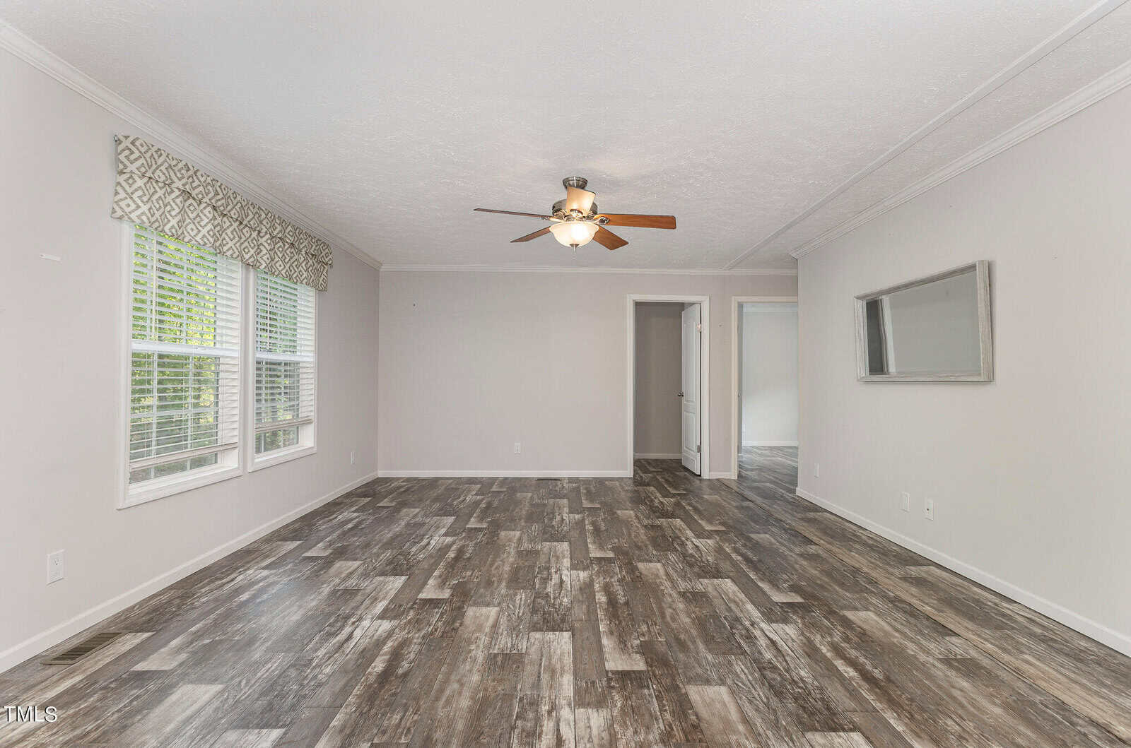 A spacious living room with gray wood flooring, light gray walls, and a ceiling fan. Large windows with patterned curtains provide natural light. A doorway leads to another room, and a rectangular mirror hangs on the wall.