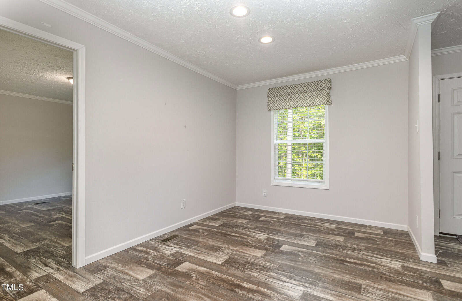 A bright, empty room with white walls and ceiling. Natural light streams through a window with a patterned valance. The floor is covered with wood-patterned tile. An open doorway leads to another room with similar flooring.