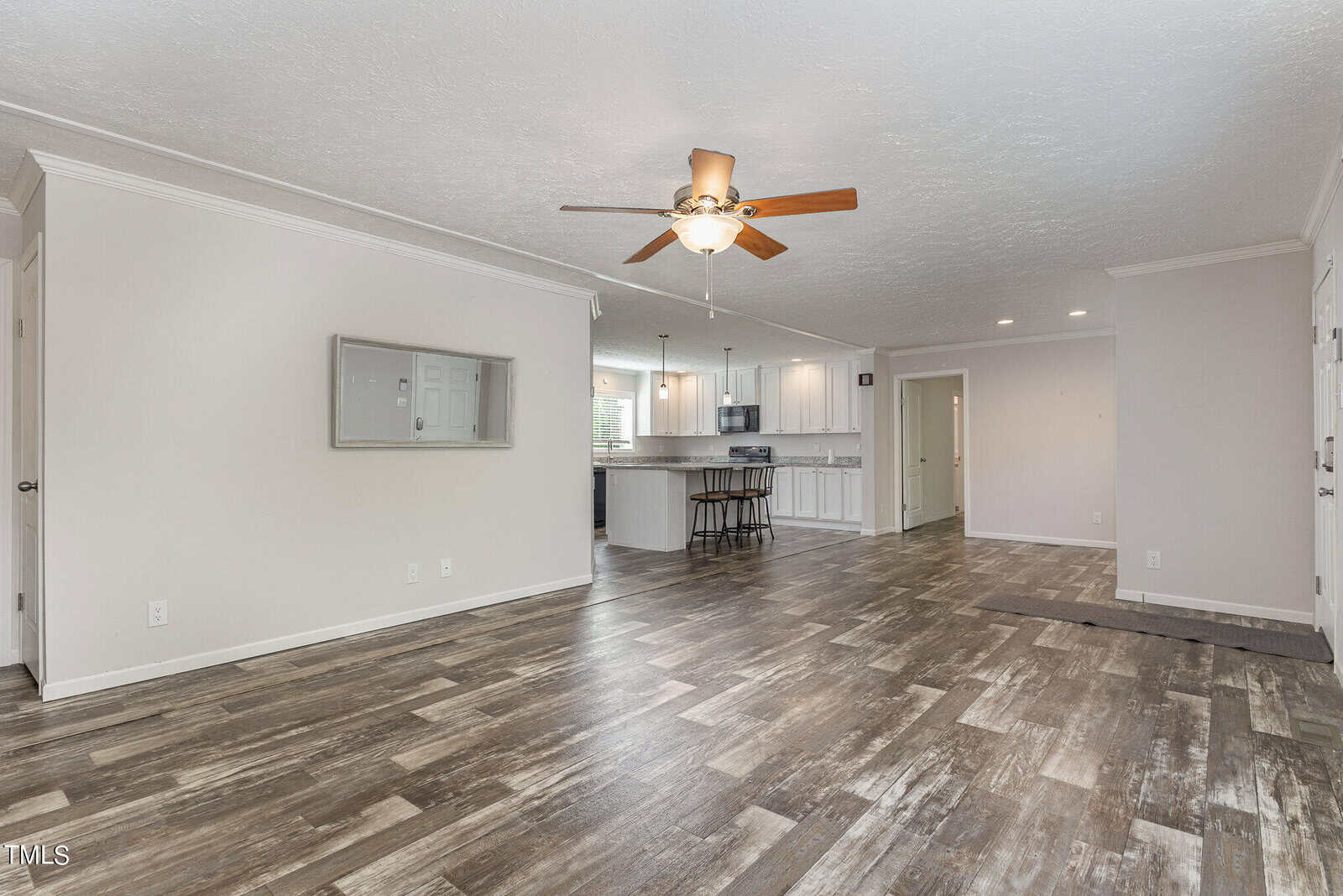 Spacious open-plan room with wood-style flooring, neutral walls, and a ceiling fan. The room includes a modern kitchen with white cabinets and a breakfast bar. Large windows offer ample natural light.