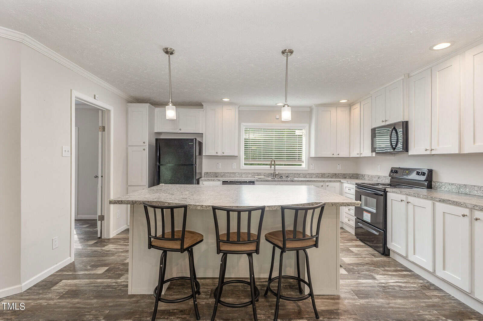 Modern kitchen with white cabinets and granite countertops. An island with three stools is centered under pendant lights. Stainless steel appliances, including a refrigerator and stove, are visible. A large window provides natural light.