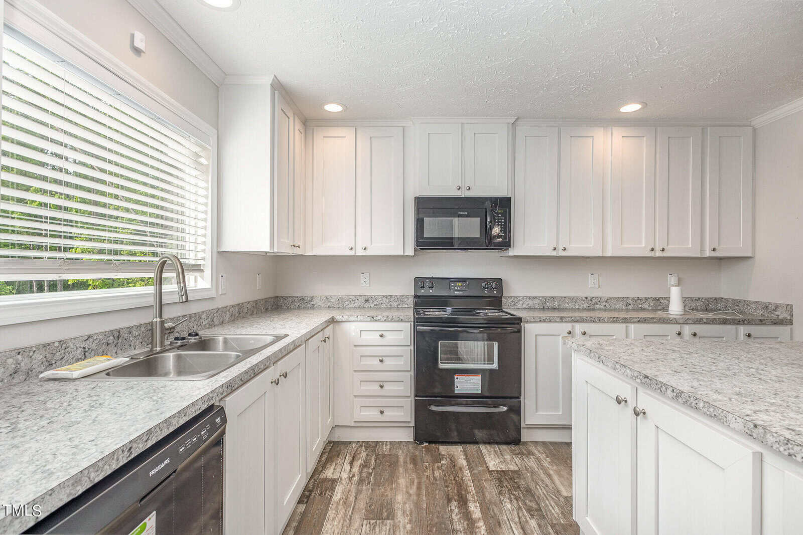 A modern kitchen with white cabinets and granite countertops. It features a black stove, microwave, and dishwasher. The kitchen has a large window over the sink and wood-patterned flooring.