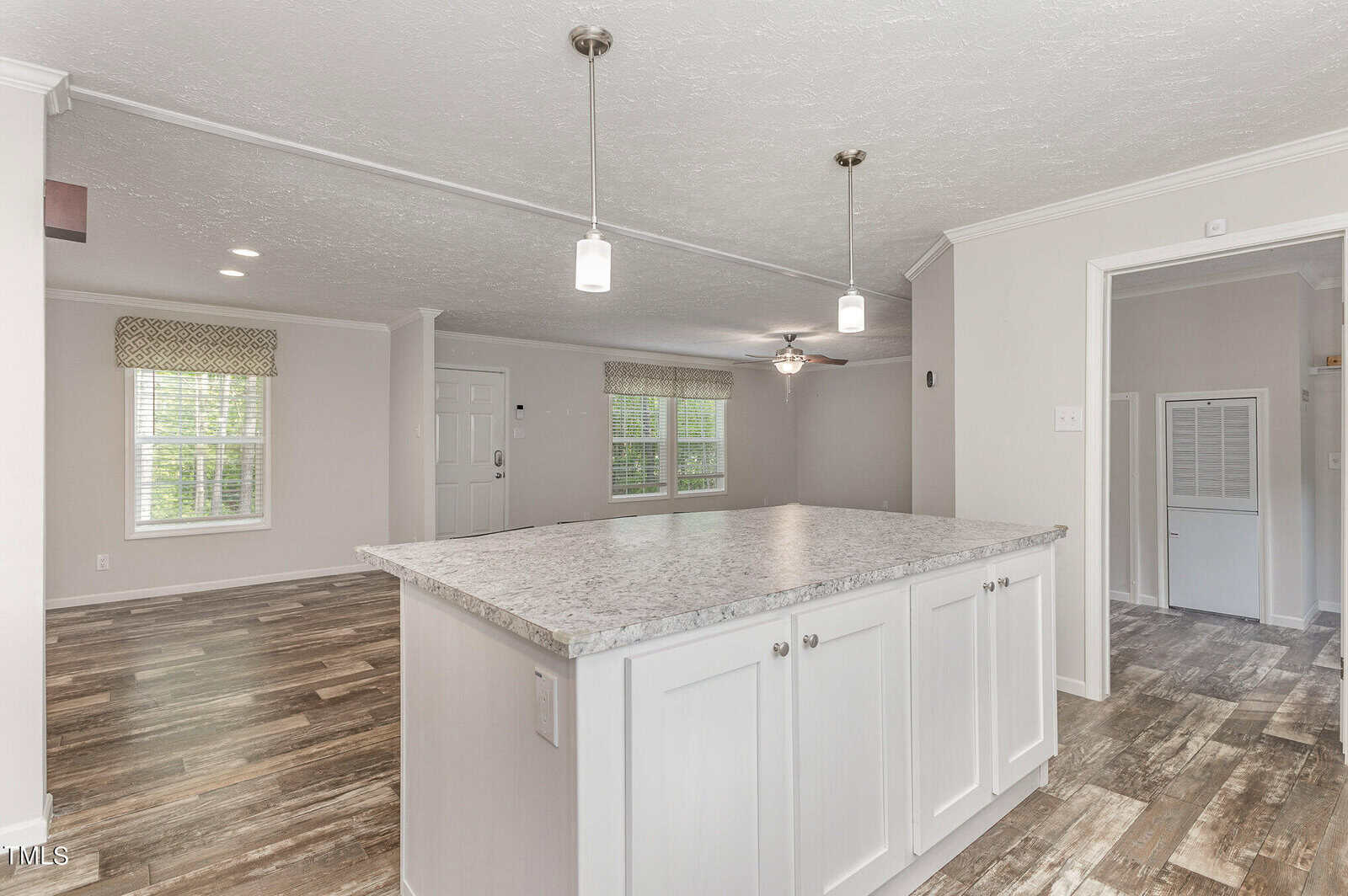 Spacious open-concept kitchen and living area featuring a large island with a marble countertop. The room has hanging pendant lights, wood-look flooring, and multiple windows with patterned valances, creating a fresh and inviting atmosphere.