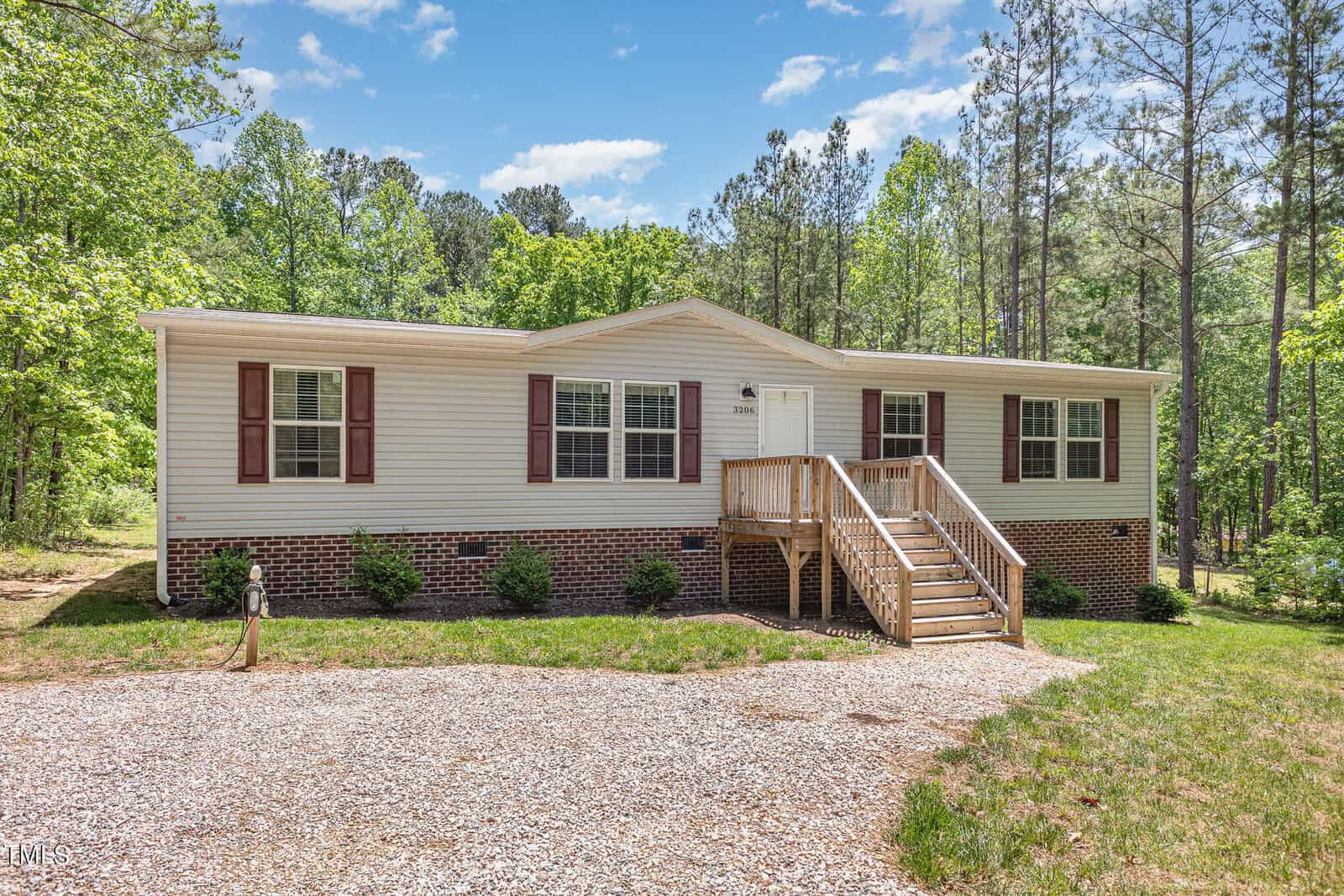 Located at 3206 Granville Ridge Road, this beige single-story house in Wake Forest features a brick foundation and brown shutters, nestled among trees. A wooden staircase leads to the front door, while a gravel driveway greets you in the foreground beneath a sky dotted with scattered clouds.