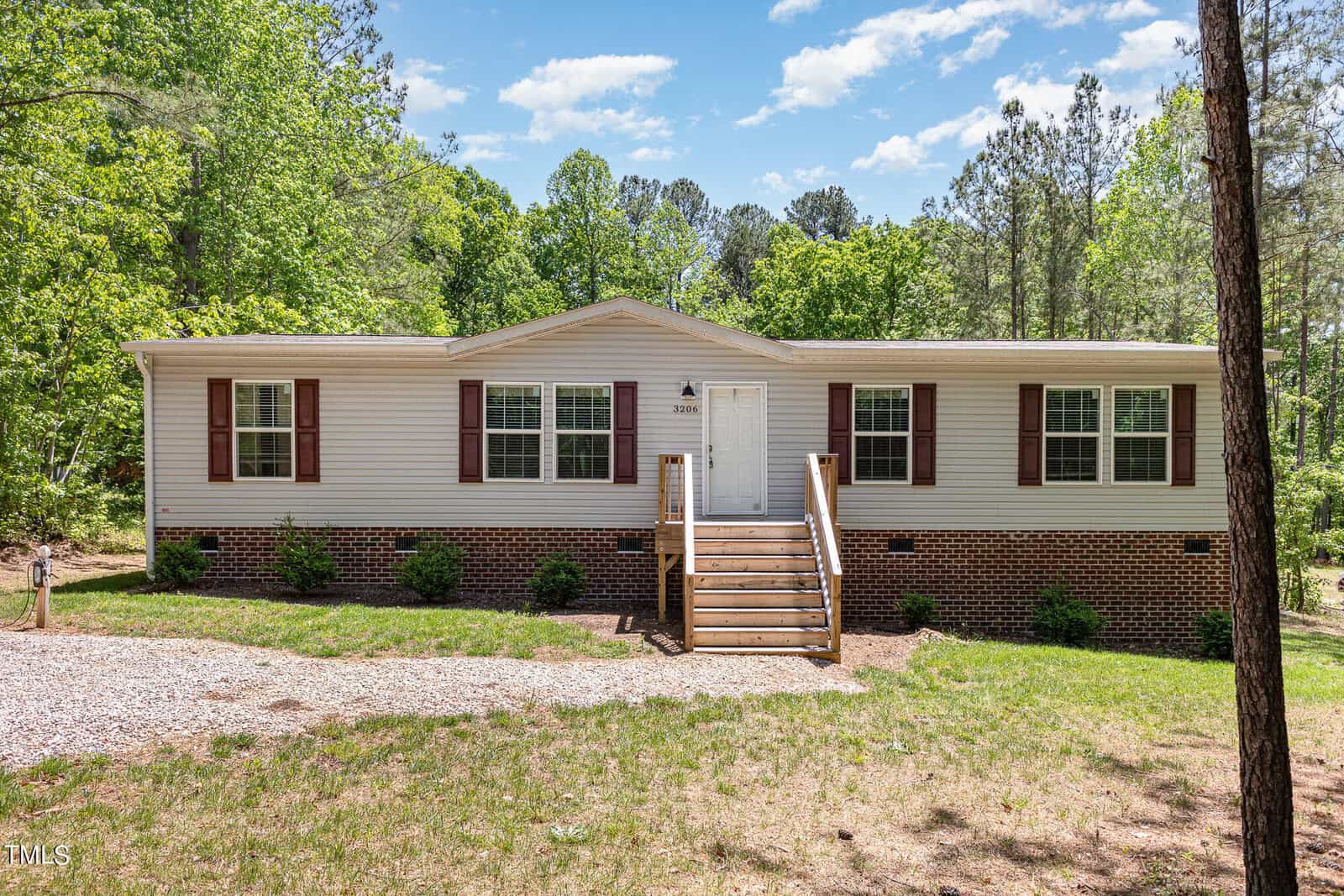 A single-story house with beige siding and a brick foundation, featuring a front porch with steps leading to a white door. It is surrounded by grass, a gravel walkway, and trees under a partly cloudy sky.
