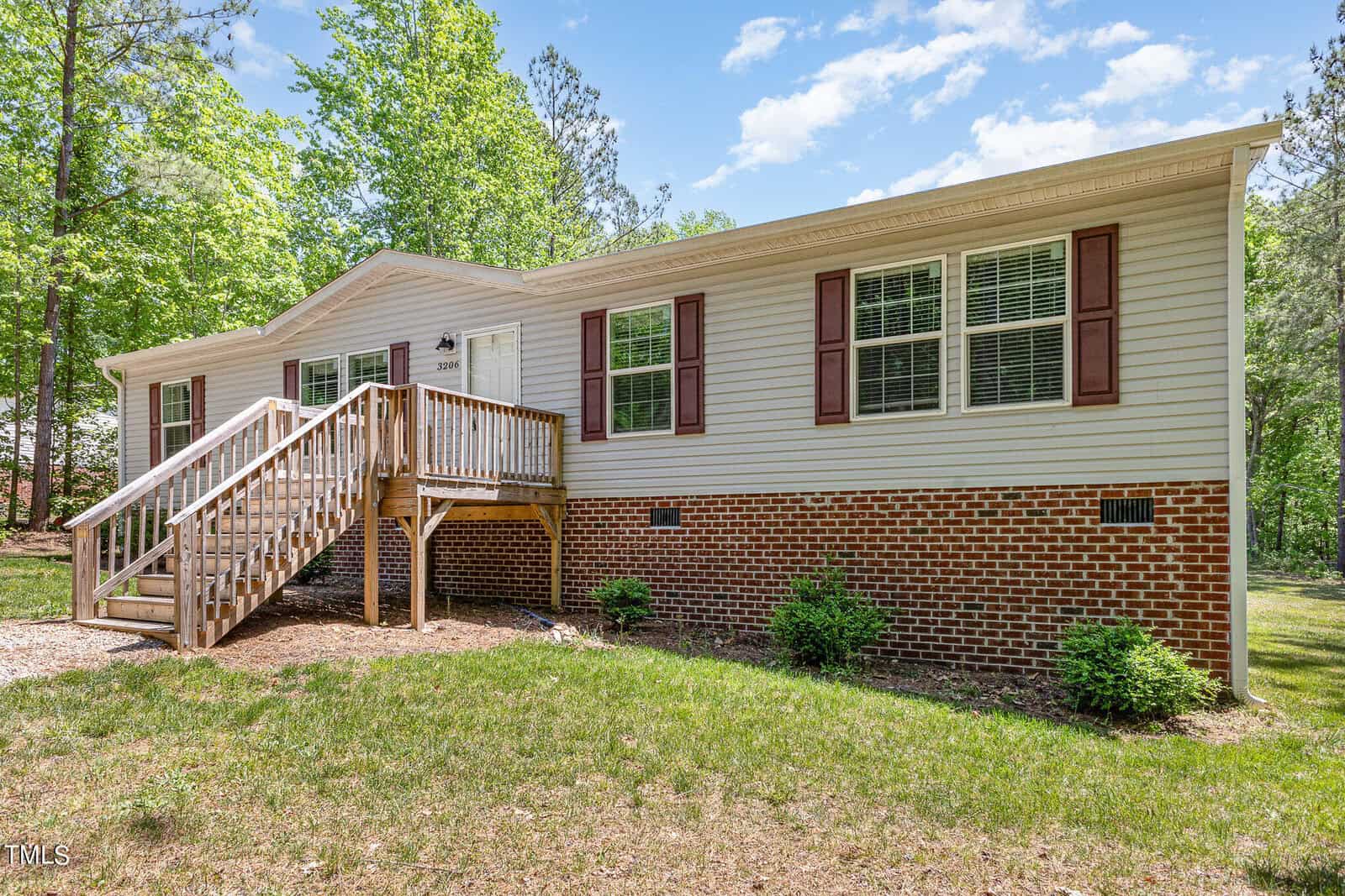 A single-story house with beige siding and a brick foundation, featuring brown shutters and a wooden front staircase. It's surrounded by grass and trees under a partly cloudy sky.