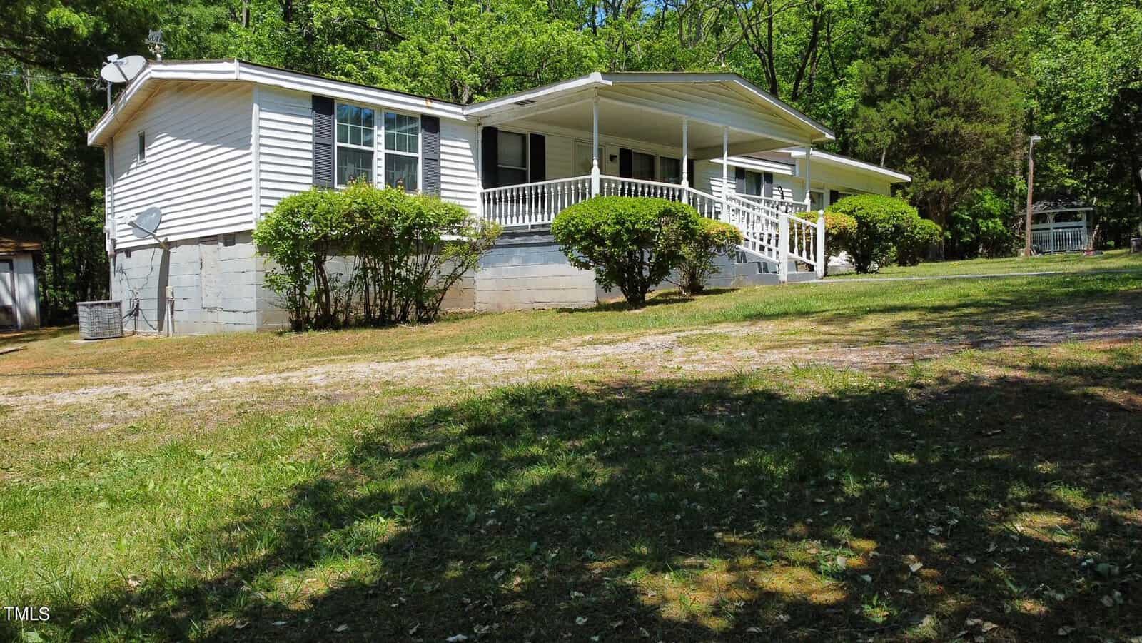 A white mobile home with a covered porch on 7701 Goshen Road is surrounded by lush greenery and bushes. Sitting on a slightly elevated foundation with steps leading up, it features a satellite dish attached to the side. The sunlit yard is grassy, adding charm to this Oxford retreat.