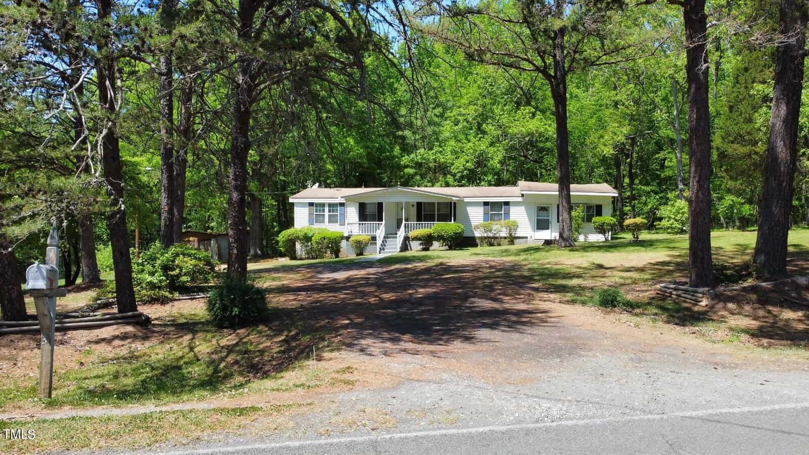A white, single-story manufactured home is nestled among tall trees. The house features a front porch with steps leading to the entrance. A paved driveway extends from the road to the home, surrounded by a grassy yard and greenery.
