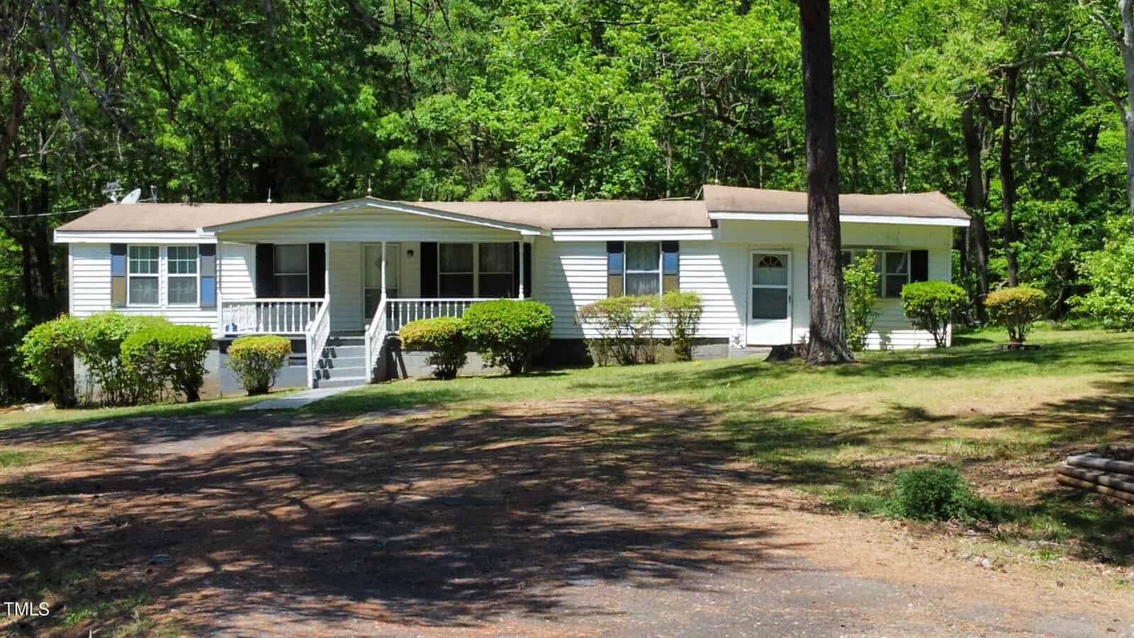 A single-story, white mobile home with a wooden front porch, surrounded by green trees and bushes. The driveway is in the foreground, leading to the house, and the scene is under sunny, clear skies.