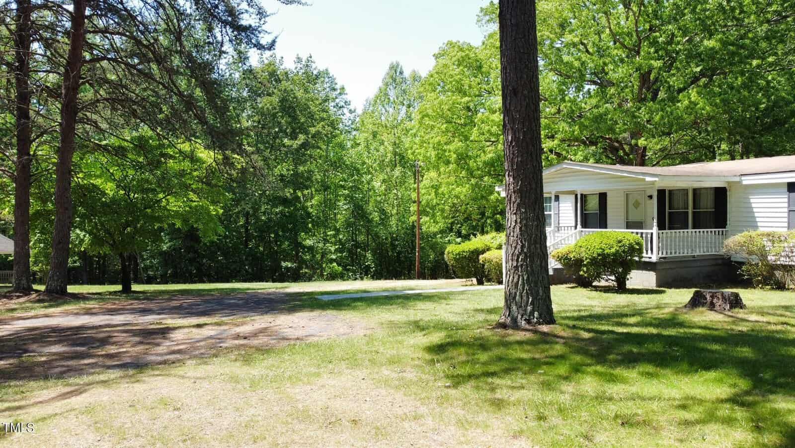A small white house with a porch sits surrounded by trees and lush greenery. A well-maintained lawn stretches in front, with a driveway to the left. Sunlight filters through the trees, casting gentle shadows on the ground.
