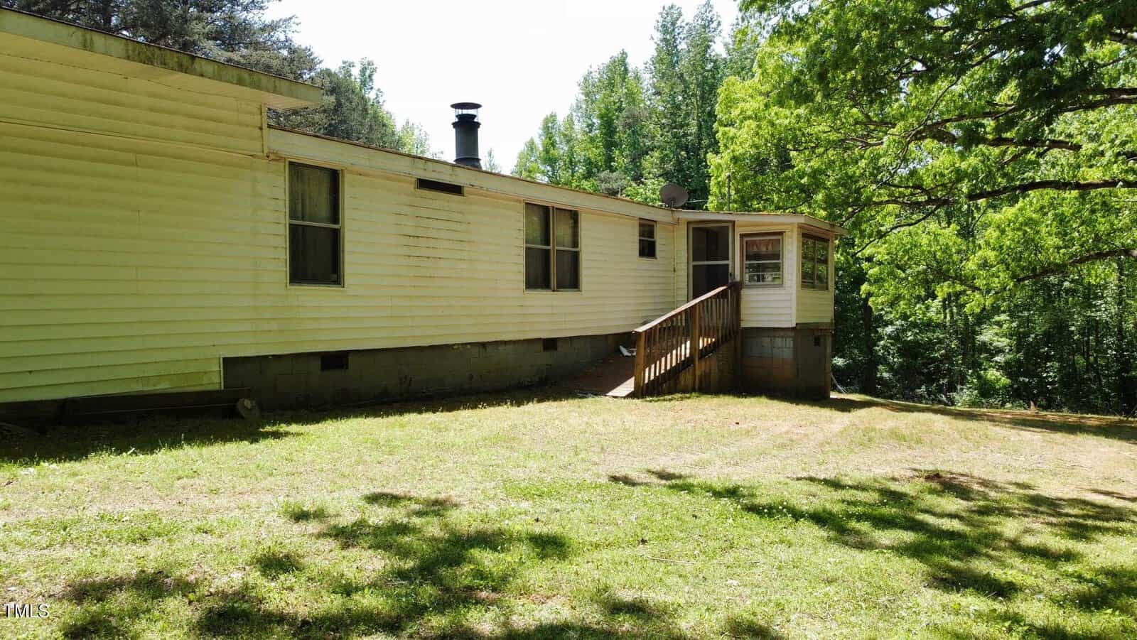 A single-story mobile home with a small porch and wooden steps, surrounded by lush green grass and trees. The house has a simple exterior with off-white siding and several windows. It's a sunny day with blue skies.