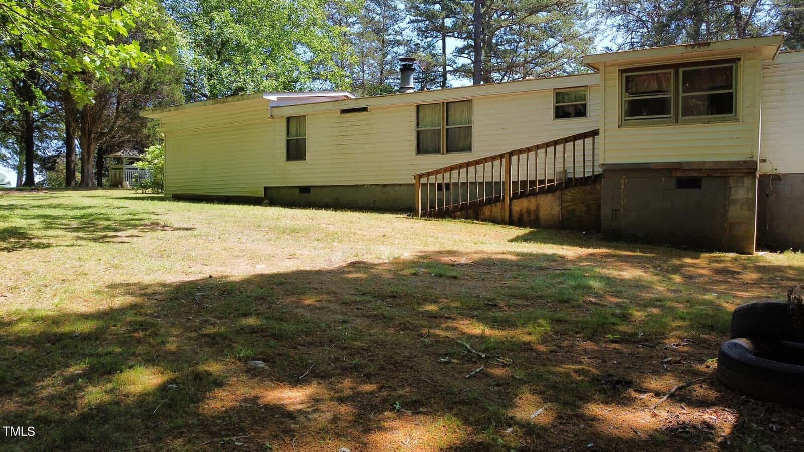 A white mobile home with a wooden ramp entrance surrounded by trees. It's set on a grassy yard with some shade from nearby trees. The foundation is elevated on concrete blocks, and there are tires stacked in the foreground.