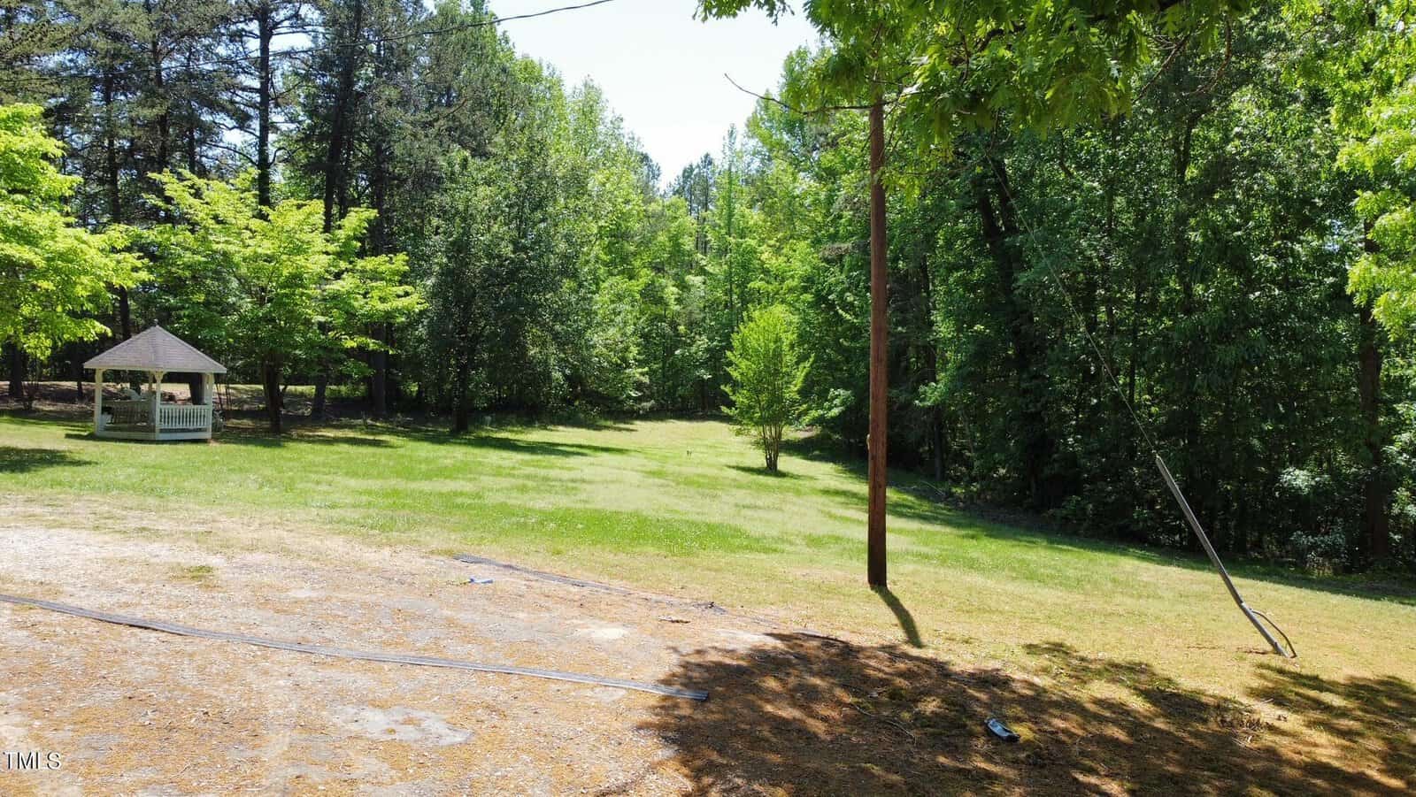 A peaceful, grassy area bordered by lush green trees. A small, white gazebo sits to the left. A utility pole with wires is on the right. Sunlight filters through the leaves, creating a tranquil atmosphere.