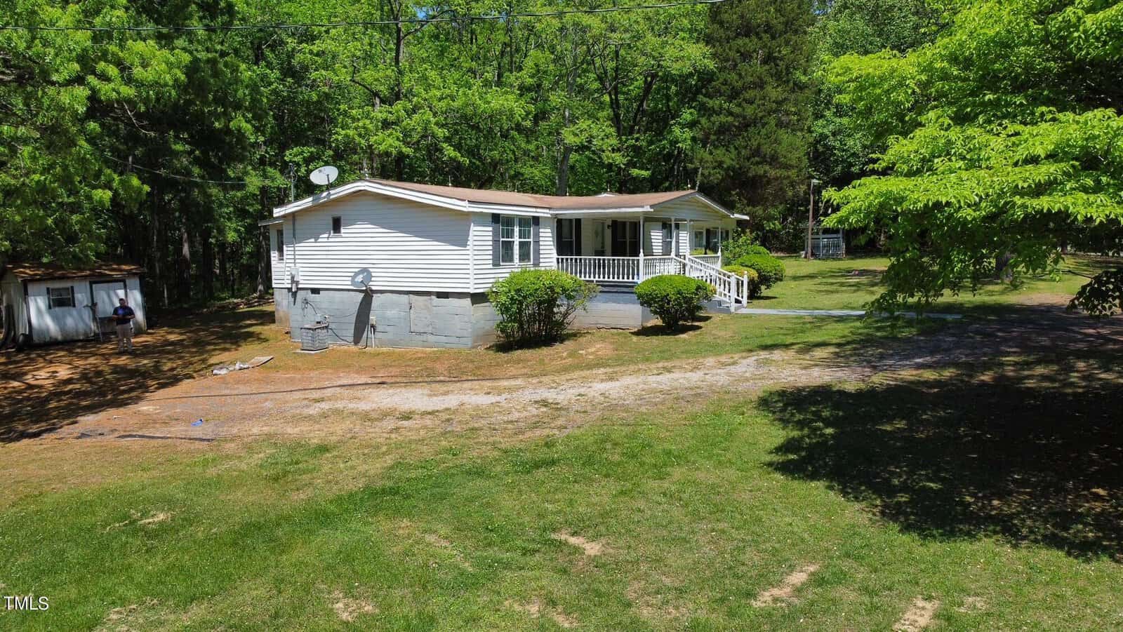 A single-story white house with a sloped roof surrounded by trees and greenery. A dirt driveway leads to the front porch. A small shed is visible in the background among the trees. The scene is sunlit and lush.