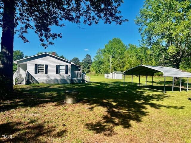 A small, single-story house with a light-colored exterior stands in a grassy yard. It has a front porch with steps. Nearby, a metal carport provides shade. Tall trees surround the property under a clear blue sky.
