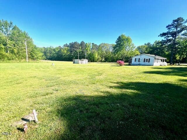 A vast grassy field under a clear blue sky, featuring a small white house and a shed in the distance. The scene is bordered by trees, and there's a cluster of pink flowers near the house.