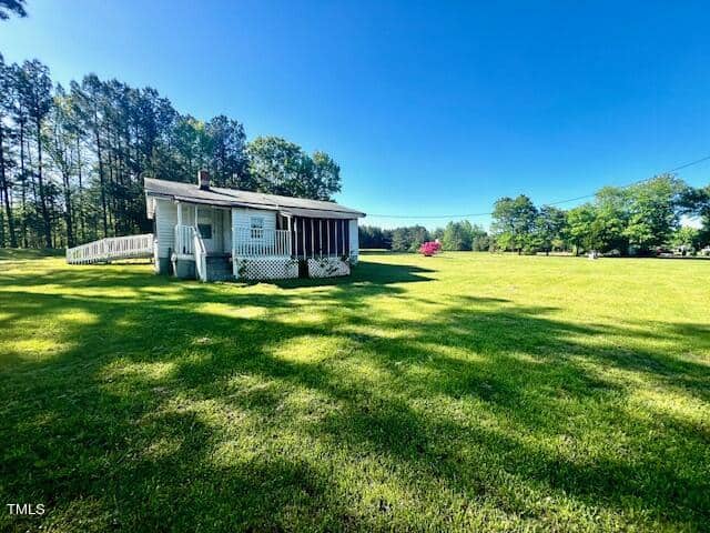 A small, white house with a ramp and porch sits on a vast, sunny green lawn. Tall trees in the background line the edge of the property under a clear blue sky.