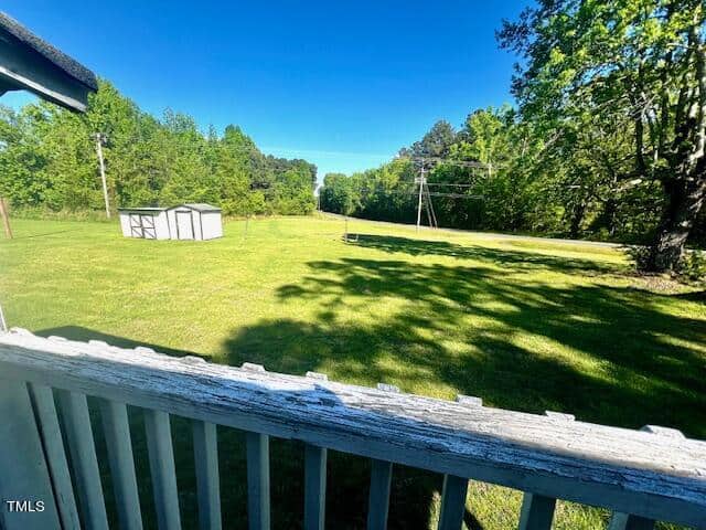 A backyard view with a wooden deck railing in the foreground, a grassy lawn, and a small white shed. Trees and power lines are visible under a clear blue sky.