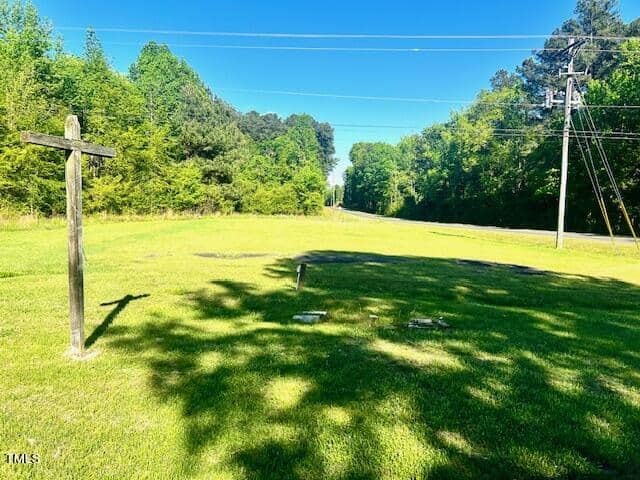 A grassy field with a wooden cross, scattered small markers, and a few patches of shade. Trees line the distant edge of the field, and a road runs alongside, bordered by utility poles and power lines.