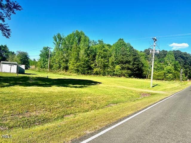 A rural scene with a paved road on the right, bordered by a grassy field and power lines. A small shed is visible on the left, and dense, leafy trees under a clear blue sky fill the background.