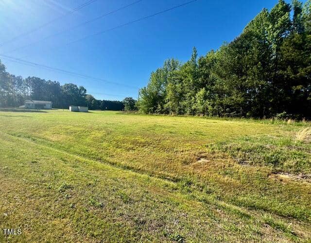Open grassy field under a clear blue sky, bordered by trees. There's a small white structure in the distance.