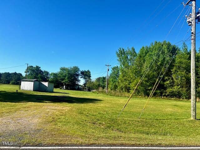 A lush green field with a few scattered trees under a clear blue sky. There are utility poles with power lines on the right and a small building in the distance to the left.