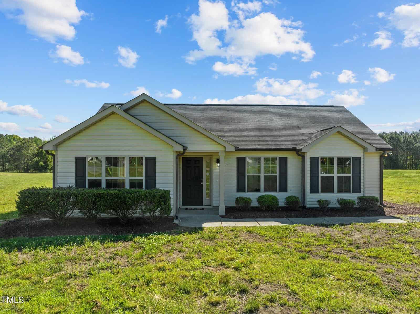 A single-story house with light siding and a dark roof sits amid a grassy field at 2210 Irvin Street under a bright blue sky with scattered clouds. Bushes line the front, and a path leads to the dark front door. Trees are visible in the background, adding to its Oxford charm.