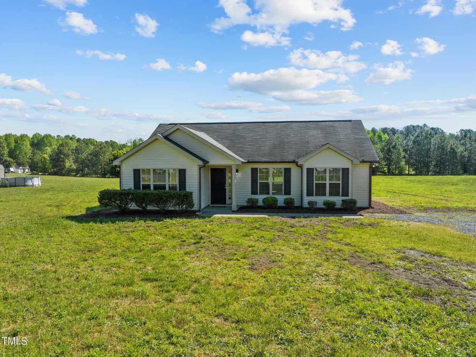A small, beige single-story house with dark shutters and a gray roof sits on a large grassy field. The sky is bright with fluffy clouds, and there are trees in the background.