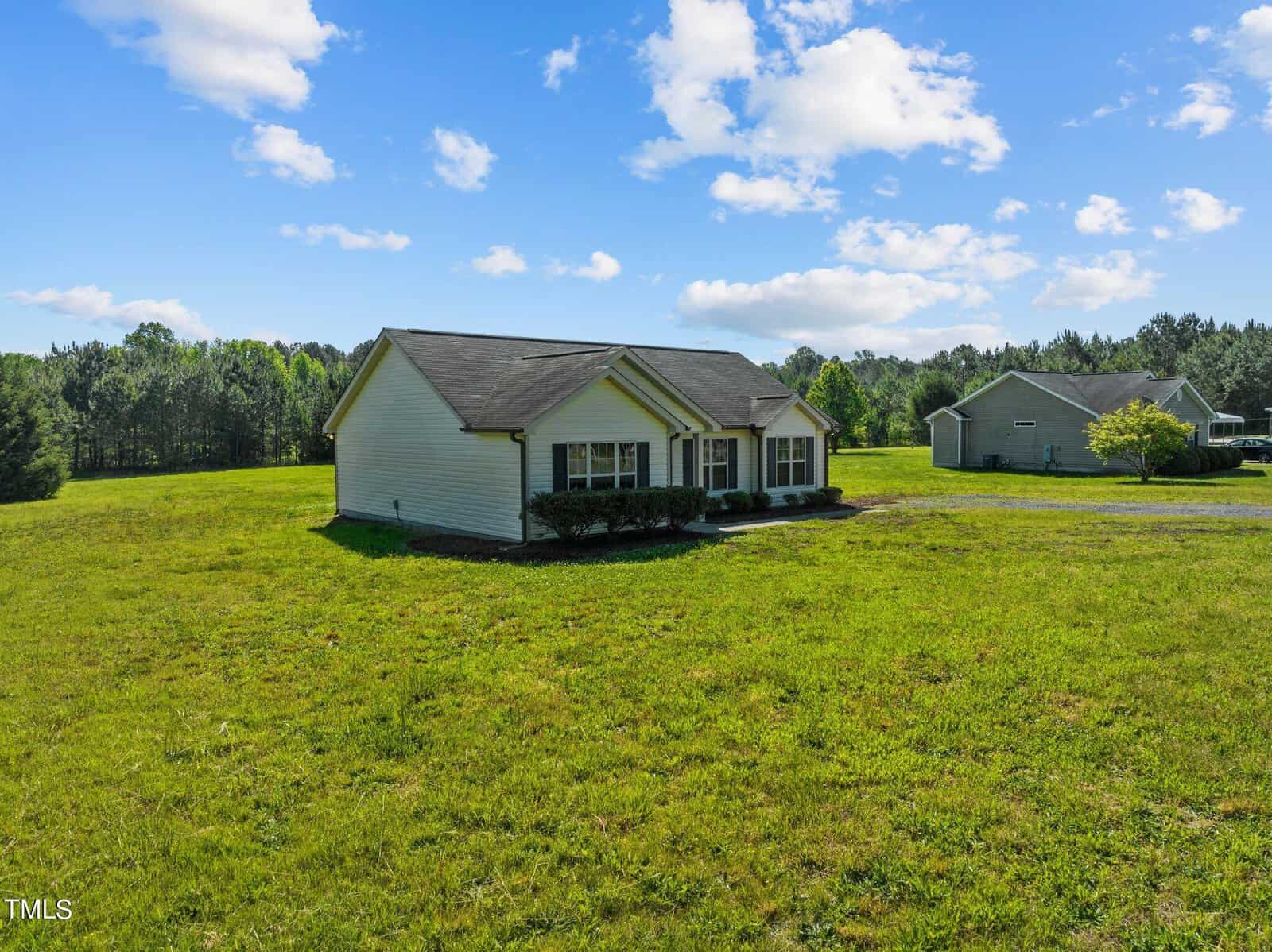 A small single-story house with a dark roof is surrounded by a vast green lawn and trees. Another house is visible in the background under a blue sky with scattered clouds.
