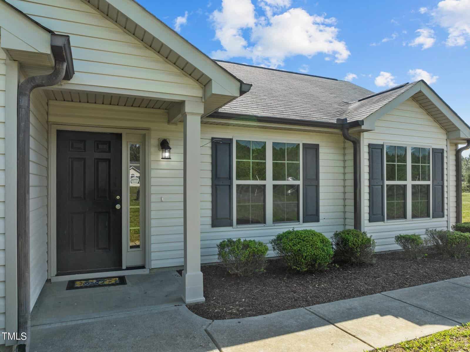 Single-story house with light-colored siding, dark shutters, and a black front door. The home features several large windows and small bushes along the walkway. A covered entryway has a light fixture near the door. The sky is partly cloudy.