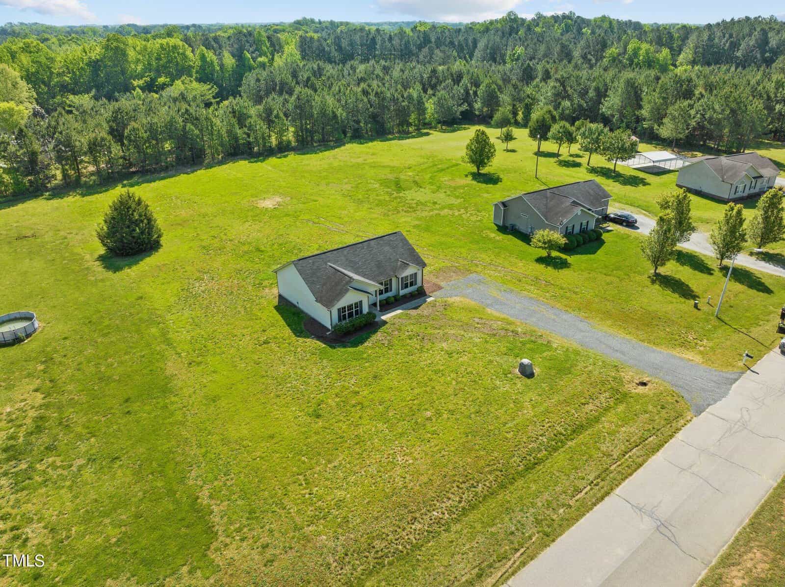 Aerial view of a rural area with scattered single-story houses on large grassy plots. A paved road runs through the neighborhood, surrounded by dense trees and open green fields under a clear blue sky.