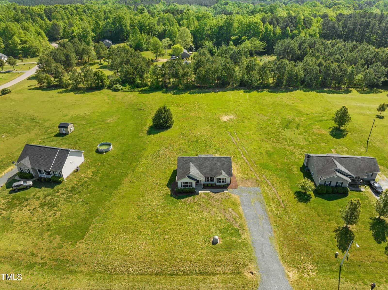 Aerial view of three single-story houses on large grassy plots with trees in the background. The central house has a driveway, and a small shed is visible near the left house. The area is surrounded by lush greenery and fields.