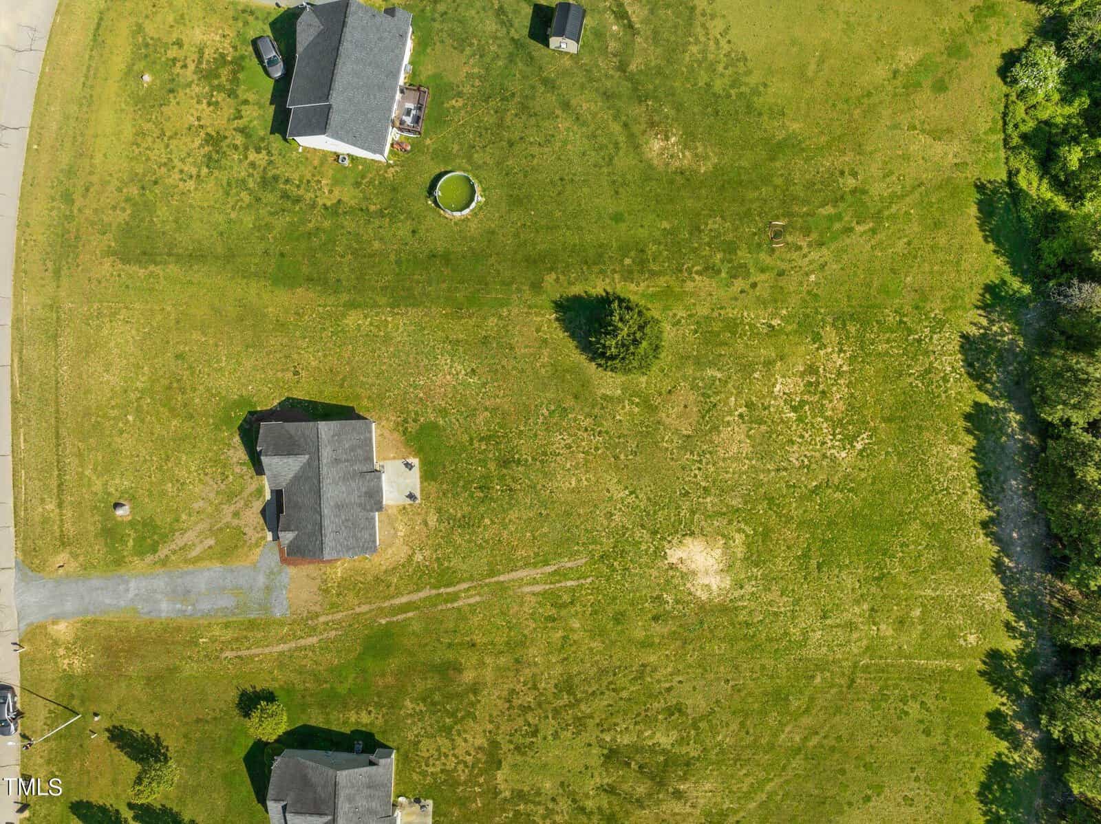 Aerial view of a suburban area with two houses on a grassy landscape, circular structure, driveway, small shed, and a patch of trees on the right side.