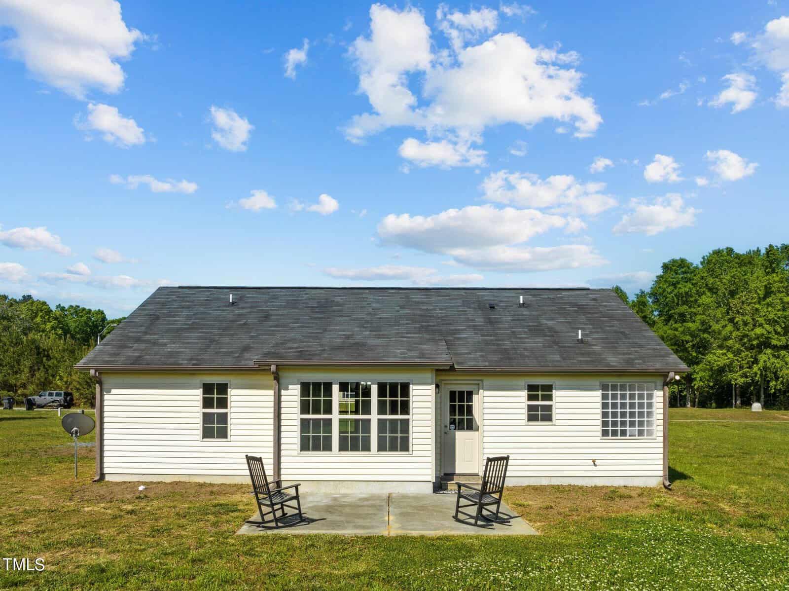 Single-story white house with a dark shingled roof, large windows, and a small concrete patio. Two rocking chairs are on the patio, set against a background of grass and trees under a partly cloudy blue sky.