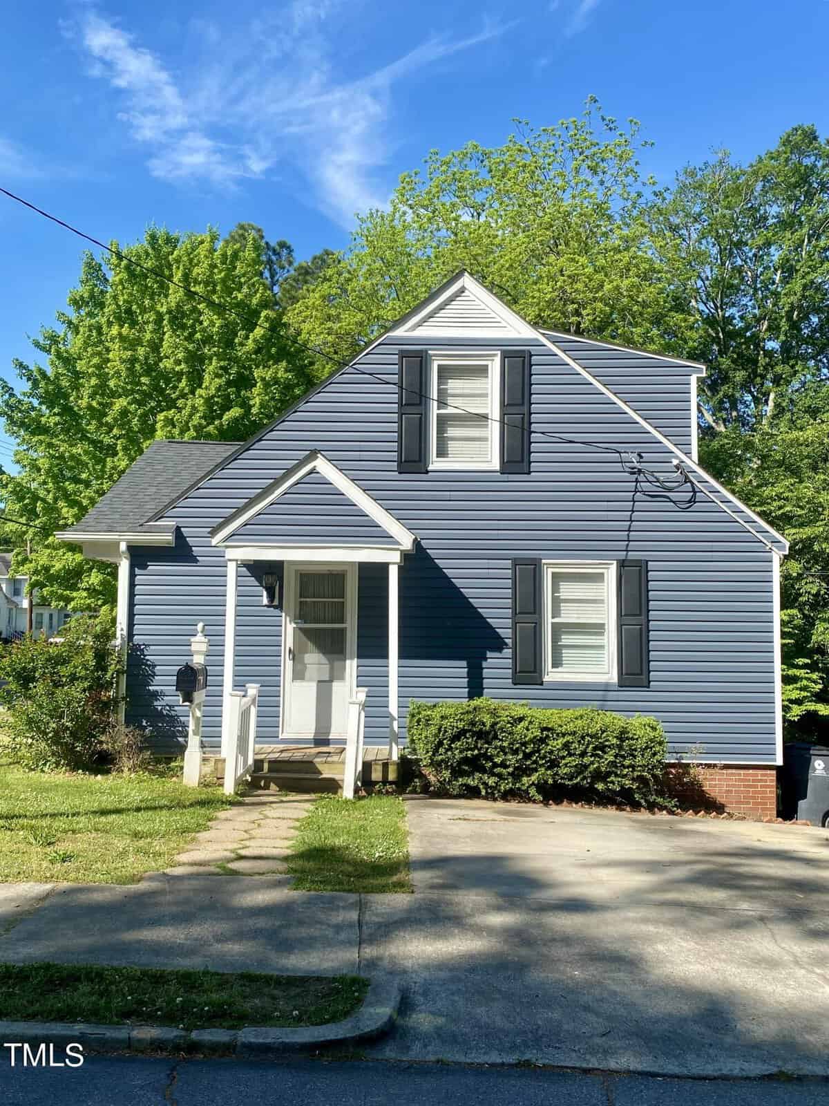 A quaint two-story blue house with white trim, featuring a small covered porch and a concrete driveway, surrounded by lush greenery under a clear blue sky.