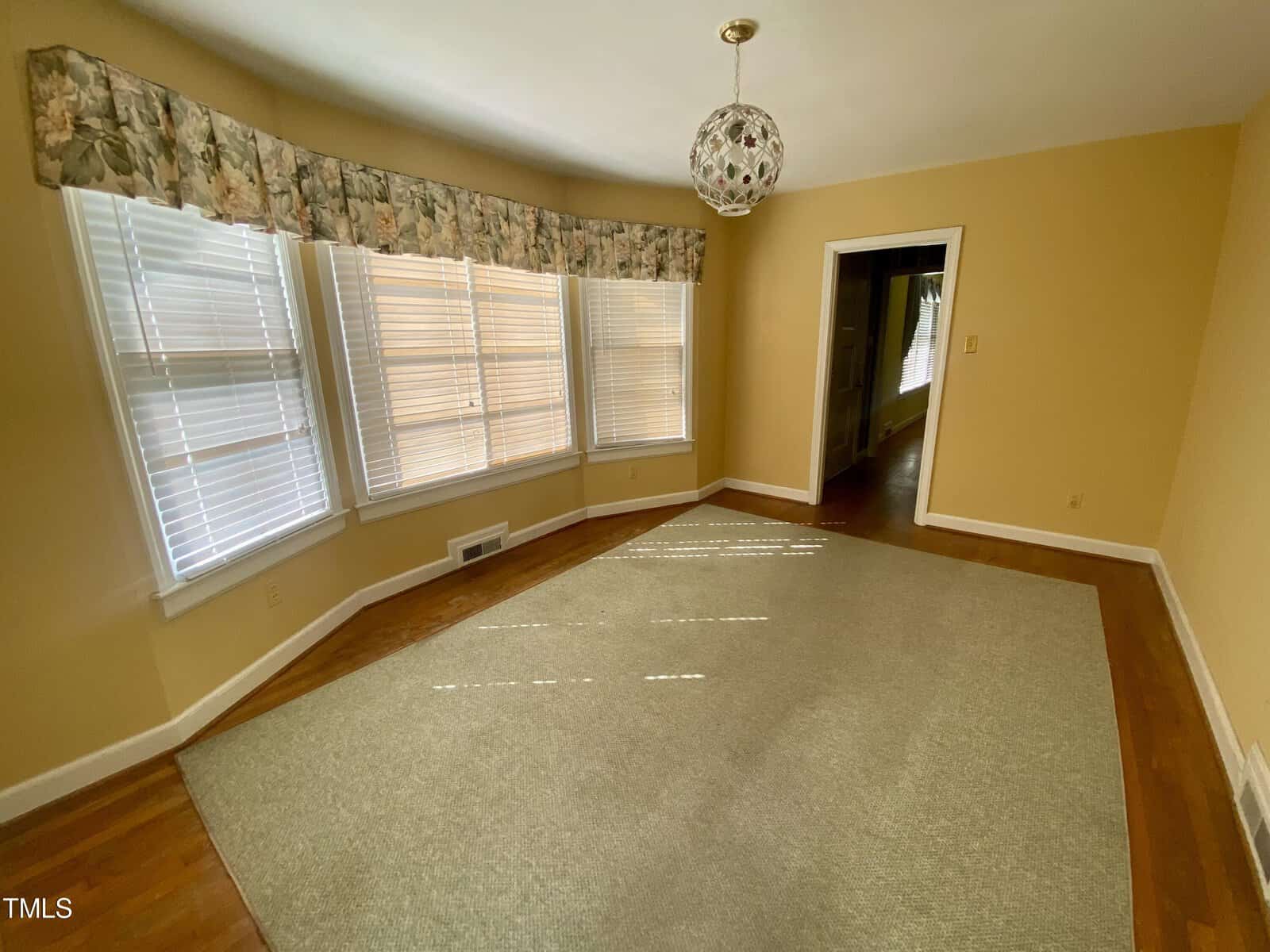 An empty room with pale yellow walls and a large window with blinds covered by a floral valance. the room has a beige carpet and a hanging glass light fixture.