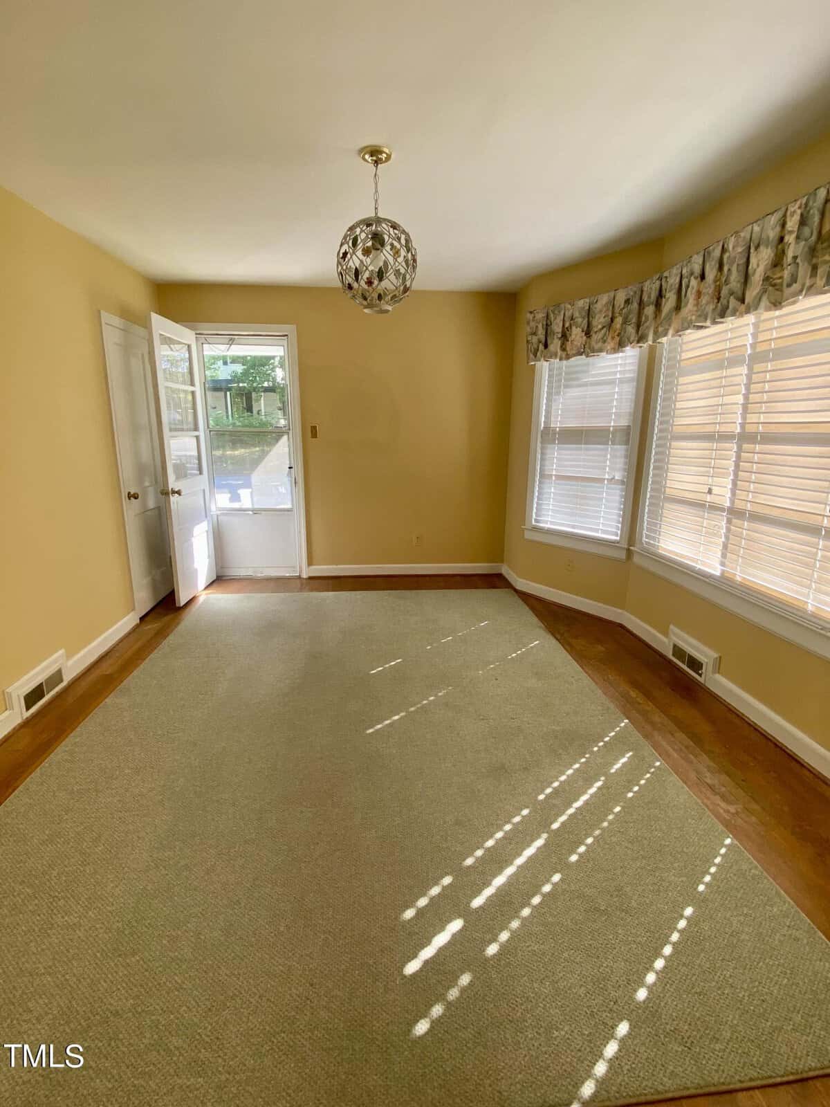 An empty room with beige carpet and light yellow walls featuring a glass door leading to a backyard, large windows with blinds, and a decorative hanging light fixture.