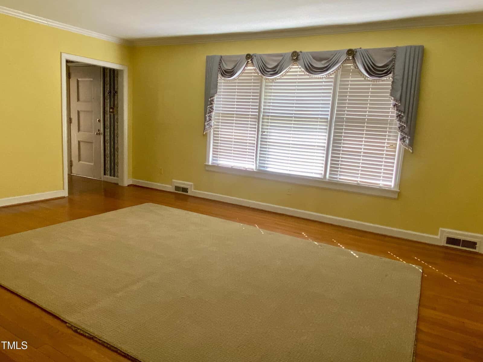 Empty living room with hardwood floors, a large beige rug, and windows dressed with grey valances. a closed door is visible on the left.