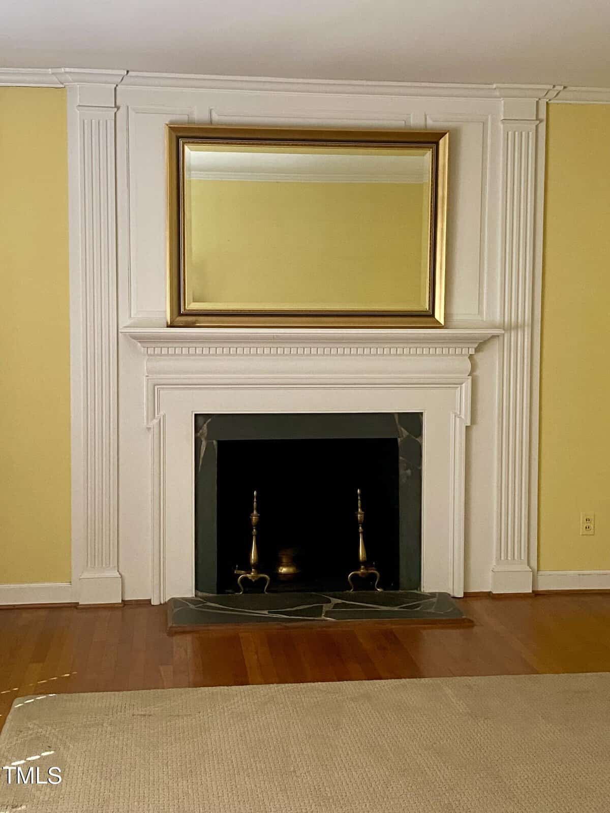 A traditional living room featuring an elegant fireplace with a white mantel, framed by cream colored walls and decorative moldings, topped with a large golden-framed mirror.