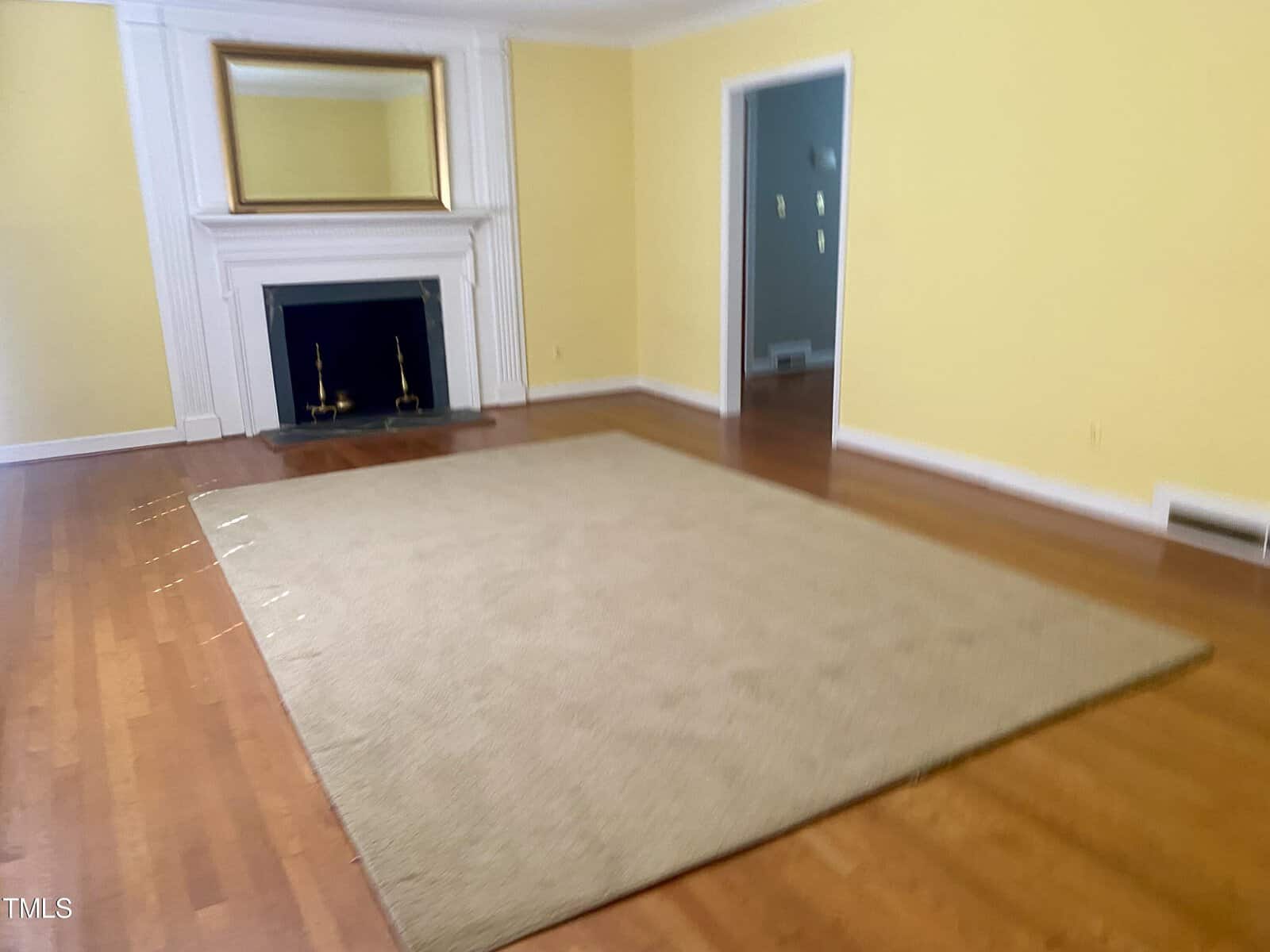 A spacious room with yellow walls, polished hardwood floors, a fireplace, and a large beige rug. two empty picture frames hang above the fireplace.