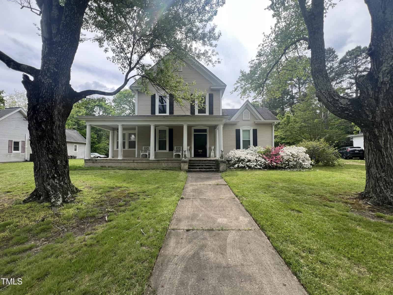 A two-story house with a porch, surrounded by lush green grass and two large trees. The front yard features a concrete pathway leading to the entrance. Flowering shrubs are near the porch, and a neighboring house is visible on the left.