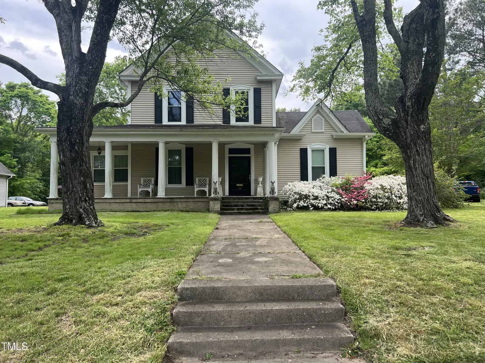 A two-story house with light-colored siding and a covered front porch, surrounded by green grass and trees. A set of steps leads up to the entrance. White flowering shrubs and a pink bush are in bloom on the right side of the house.