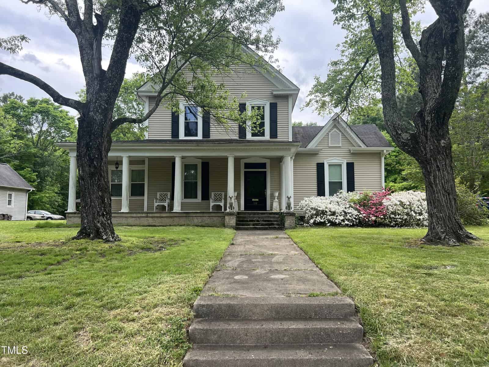 Nestled at 512 Raleigh Street, this two-story Oxford-style house boasts white siding, a covered front porch, and three large windows. It's enveloped by blooming bushes and trees, with a neatly trimmed lawn leading the way from the street via a concrete path.