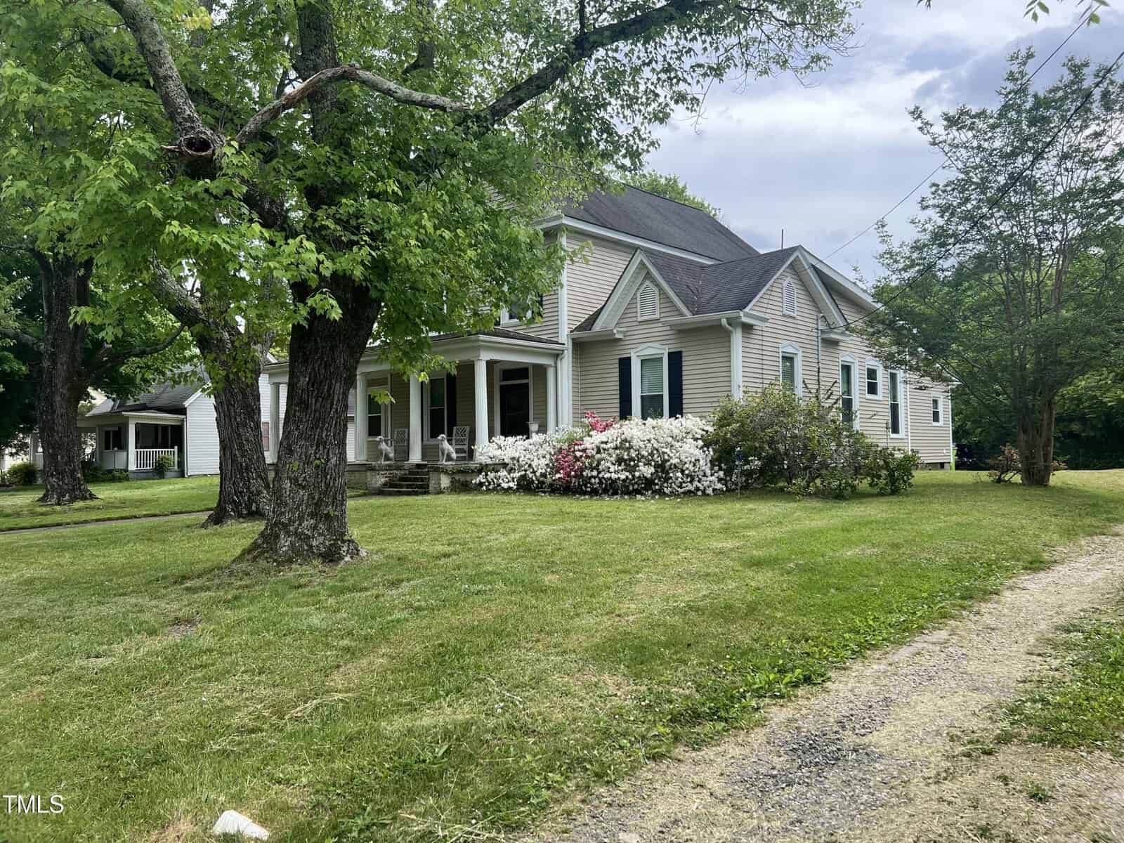A charming two-story house with light gray siding is surrounded by lush green grass and flowering bushes. Large trees provide shade to the property. A gravel path leads up to the front of the house, which features a covered porch.
